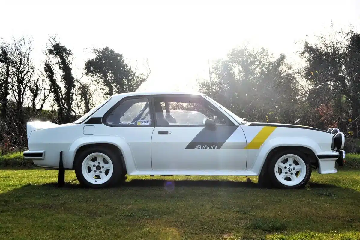 Weißer Opel Ascona 400R von 1980 im Rallye-Trim auf einer Wiese im Sonnenlicht, seitlich fotografiert.