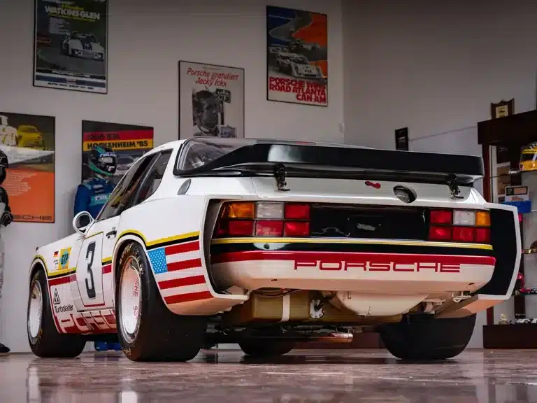 Heckansicht eines weißen Porsche 924 GTP mit Rennlackierung in einer Garage, umgeben von Motorsportpostern.