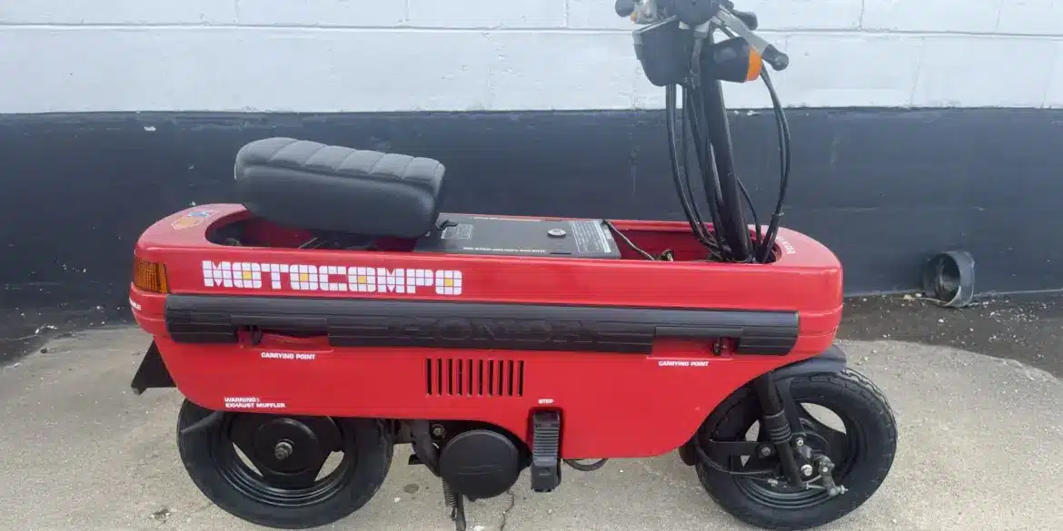 Roter Honda Motocompo Roller von 1982 im aufgeklappten Zustand vor einer weißen Wand, mit sichtbaren Lenker- und Sitzdetails.