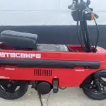 Roter Honda Motocompo Roller von 1982 im aufgeklappten Zustand vor einer weißen Wand, mit sichtbaren Lenker- und Sitzdetails.