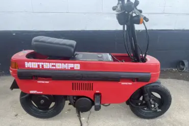 Roter Honda Motocompo Roller von 1982 im aufgeklappten Zustand vor einer weißen Wand, mit sichtbaren Lenker- und Sitzdetails.