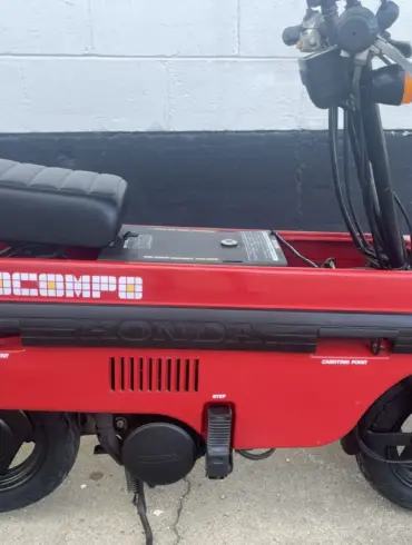 Roter Honda Motocompo Roller von 1982 im aufgeklappten Zustand vor einer weißen Wand, mit sichtbaren Lenker- und Sitzdetails.