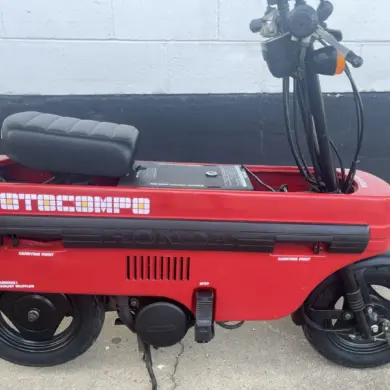 Roter Honda Motocompo Roller von 1982 im aufgeklappten Zustand vor einer weißen Wand, mit sichtbaren Lenker- und Sitzdetails.