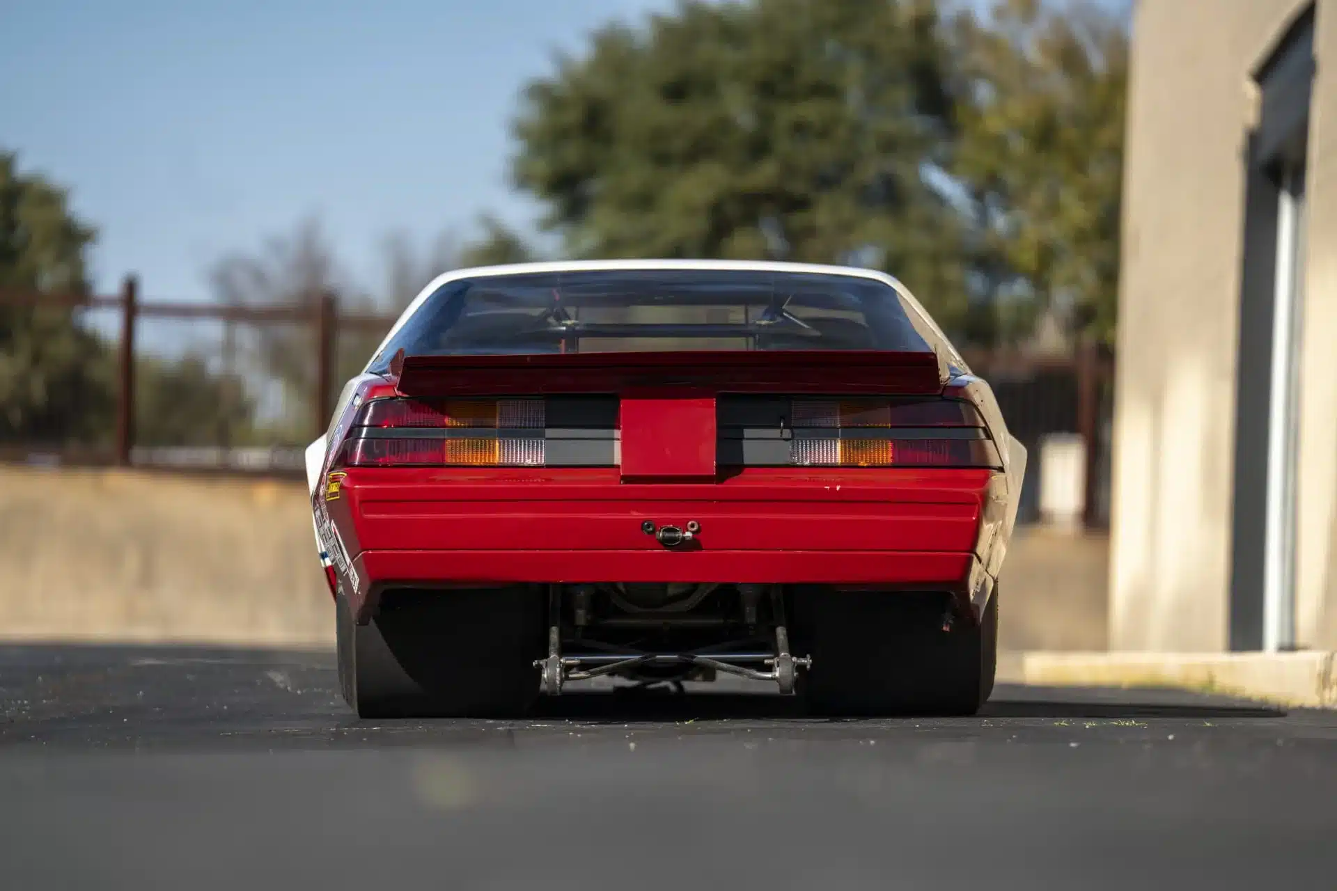 Heckansicht eines roten 1984 Chevrolet Camaro Rennwagens mit breiten Reifen und großem Spoiler, aufgenommen auf einer asphaltierten Fläche.