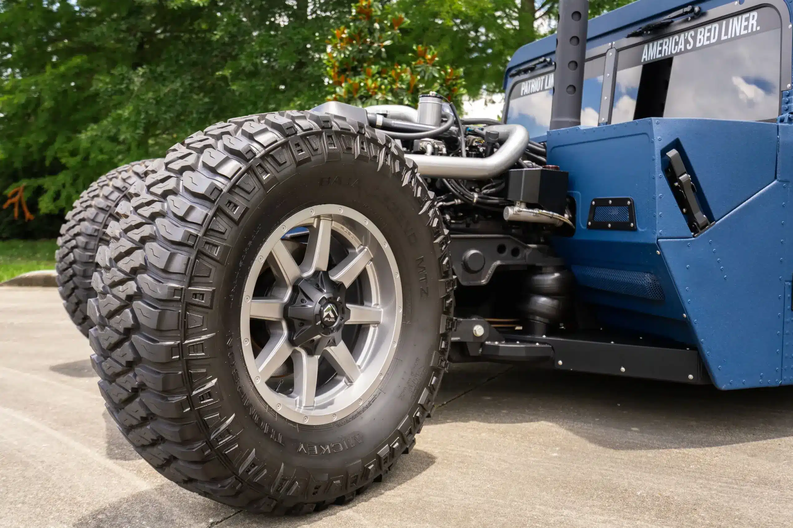 Großes Offroad-Rad mit Mickey Thompson Reifen und blauer, beschichteter Karosserie eines Humvee Restomods, Detailaufnahme mit sichtbarer Motor- und Fahrwerkskomponente.