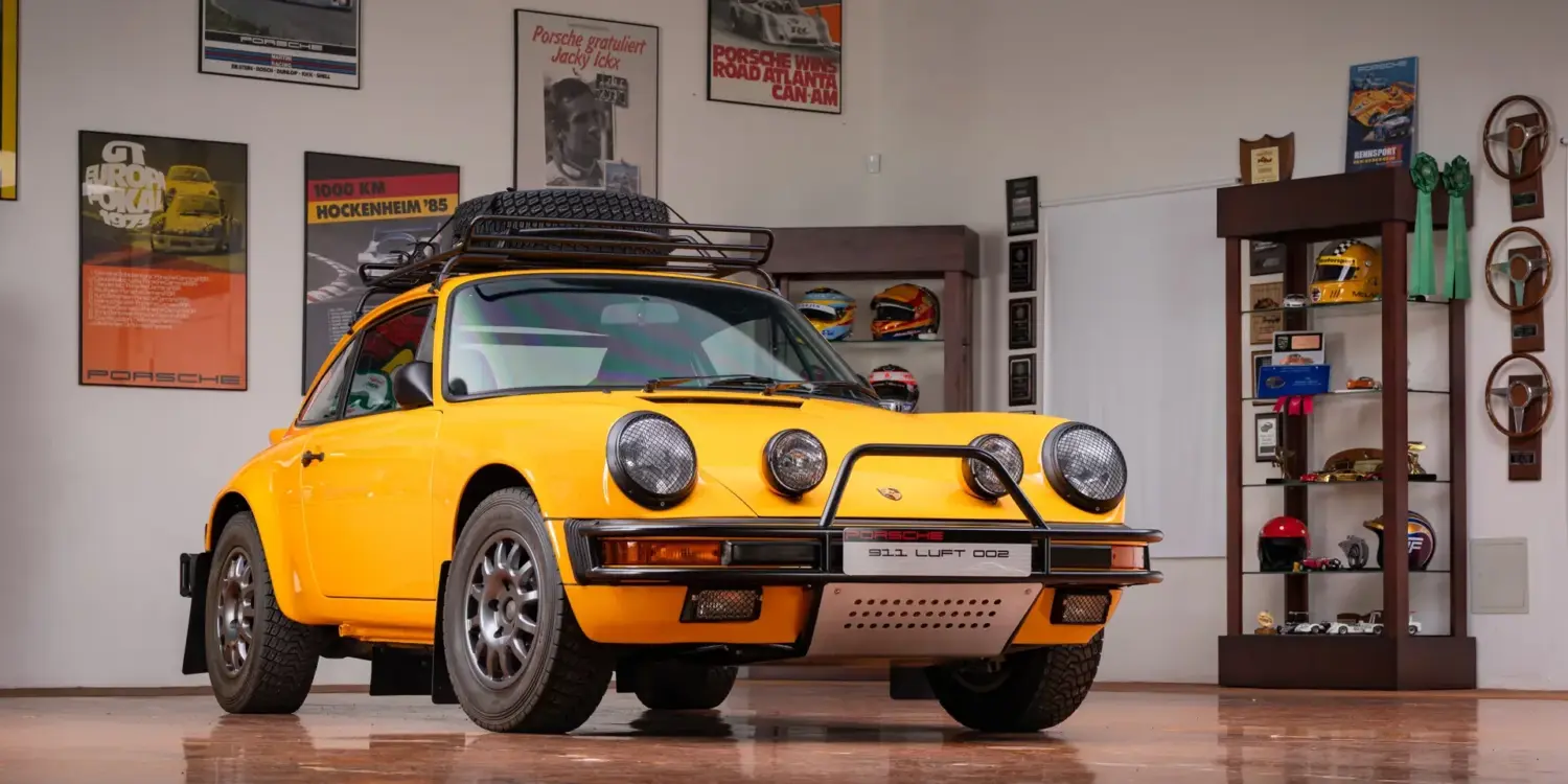 Gelber Porsche 911 Safari Umbau Baujahr 1987 mit Dachgepäckträger und Zusatzscheinwerfern in einer Garage, umgeben von Motorsport-Postern und Trophäen.