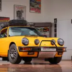 Gelber Porsche 911 Safari Umbau Baujahr 1987 mit Dachgepäckträger und Zusatzscheinwerfern in einer Garage, umgeben von Motorsport-Postern und Trophäen.