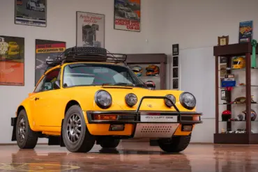 Gelber Porsche 911 Safari Umbau Baujahr 1987 mit Dachgepäckträger und Zusatzscheinwerfern in einer Garage, umgeben von Motorsport-Postern und Trophäen.