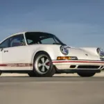 Weißer Porsche 911 Carrera Classic Study von Singer aus dem Jahr 1990 auf freier Fläche, Seitenansicht, dezente rote Akzente.