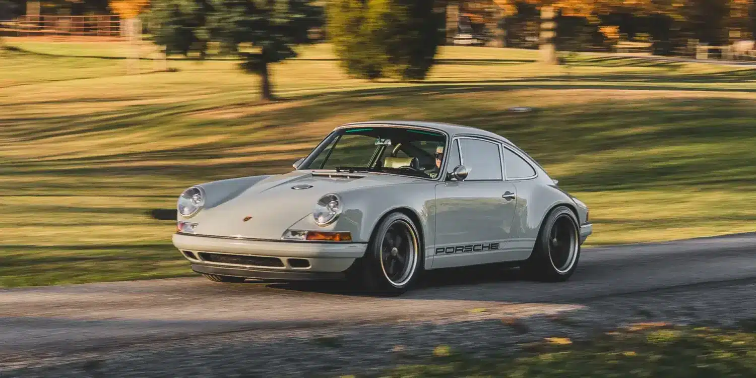 Ein Singer Reimagined Porsche 911 Novato-Commission in Dolphin Grey fährt auf einer Landstraße, umgeben von Bäumen und Wiese.