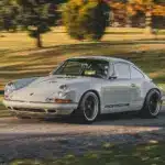 Ein Singer Reimagined Porsche 911 Novato-Commission in Dolphin Grey fährt auf einer Landstraße, umgeben von Bäumen und Wiese.