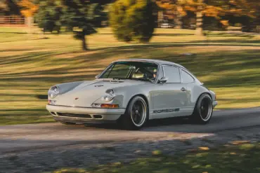 Ein Singer Reimagined Porsche 911 Novato-Commission in Dolphin Grey fährt auf einer Landstraße, umgeben von Bäumen und Wiese.