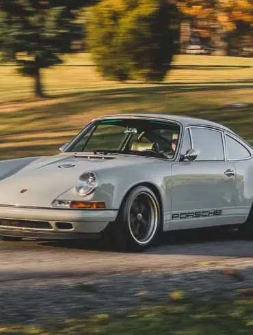 Ein Singer Reimagined Porsche 911 Novato-Commission in Dolphin Grey fährt auf einer Landstraße, umgeben von Bäumen und Wiese.