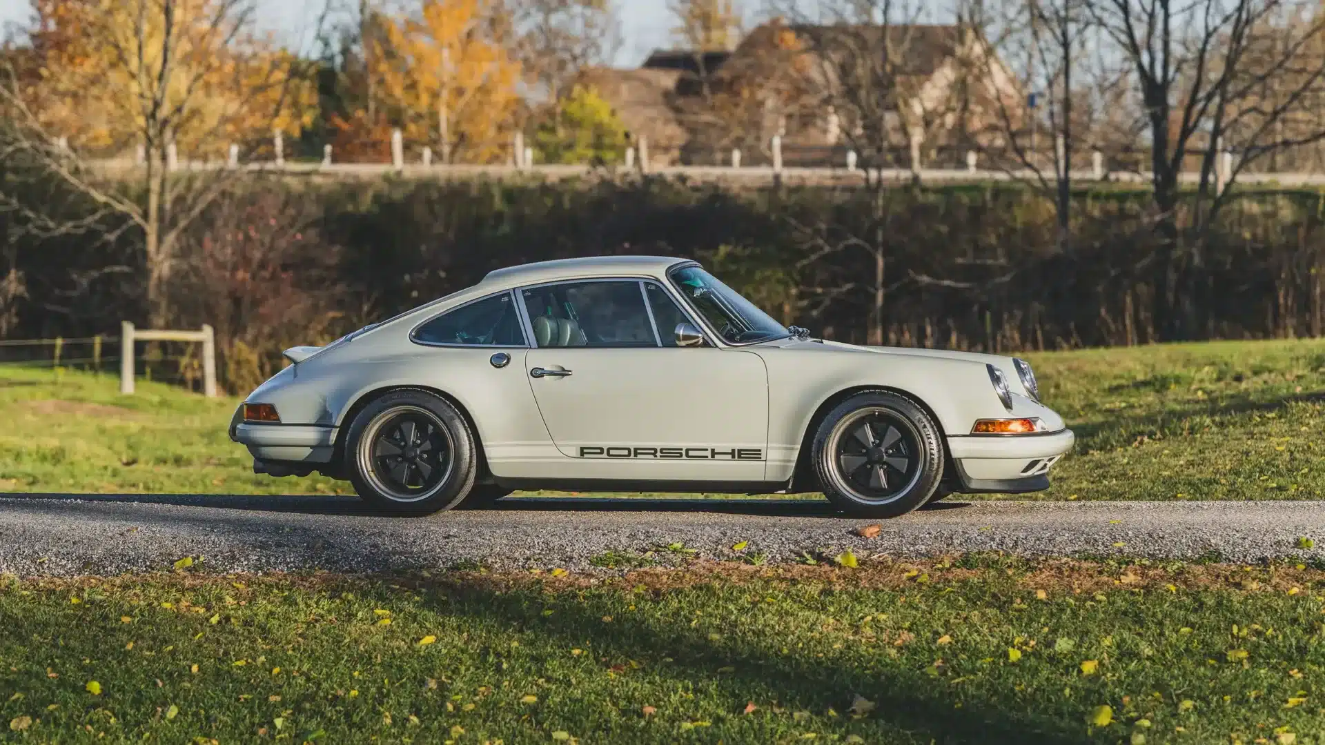 Seitenansicht eines grau lackierten Singer Porsche 911 Novato-Commission mit dunkelgrünem Porsche-Schriftzug, stehend auf einer Straße vor herbstlicher Landschaft.