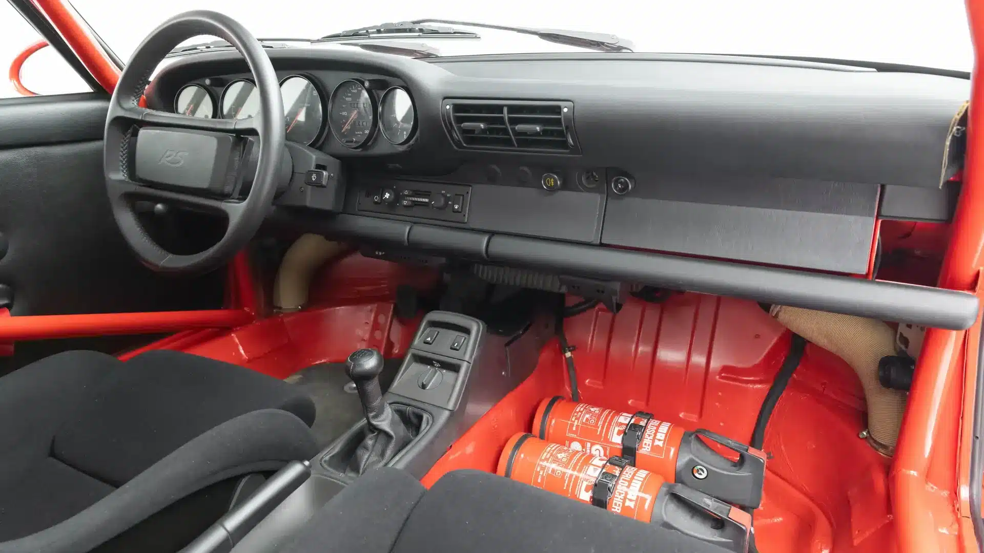 Innenraum eines Porsche 911 Carrera RS N/GT mit Rennsportsitzen, Überrollbügel und Feuerlöschern, minimalistische Ausstattung.