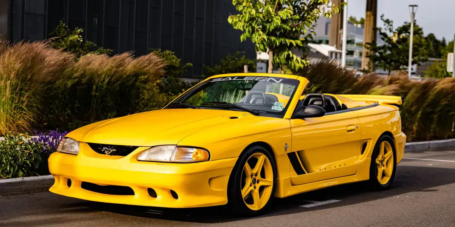 Gelber 1995 Saleen Mustang S351 Cabrio Supercharged auf einem Parkplatz vor moderner Kulisse.