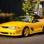 Gelber 1995 Saleen Mustang S351 Cabrio Supercharged auf einem Parkplatz vor moderner Kulisse.