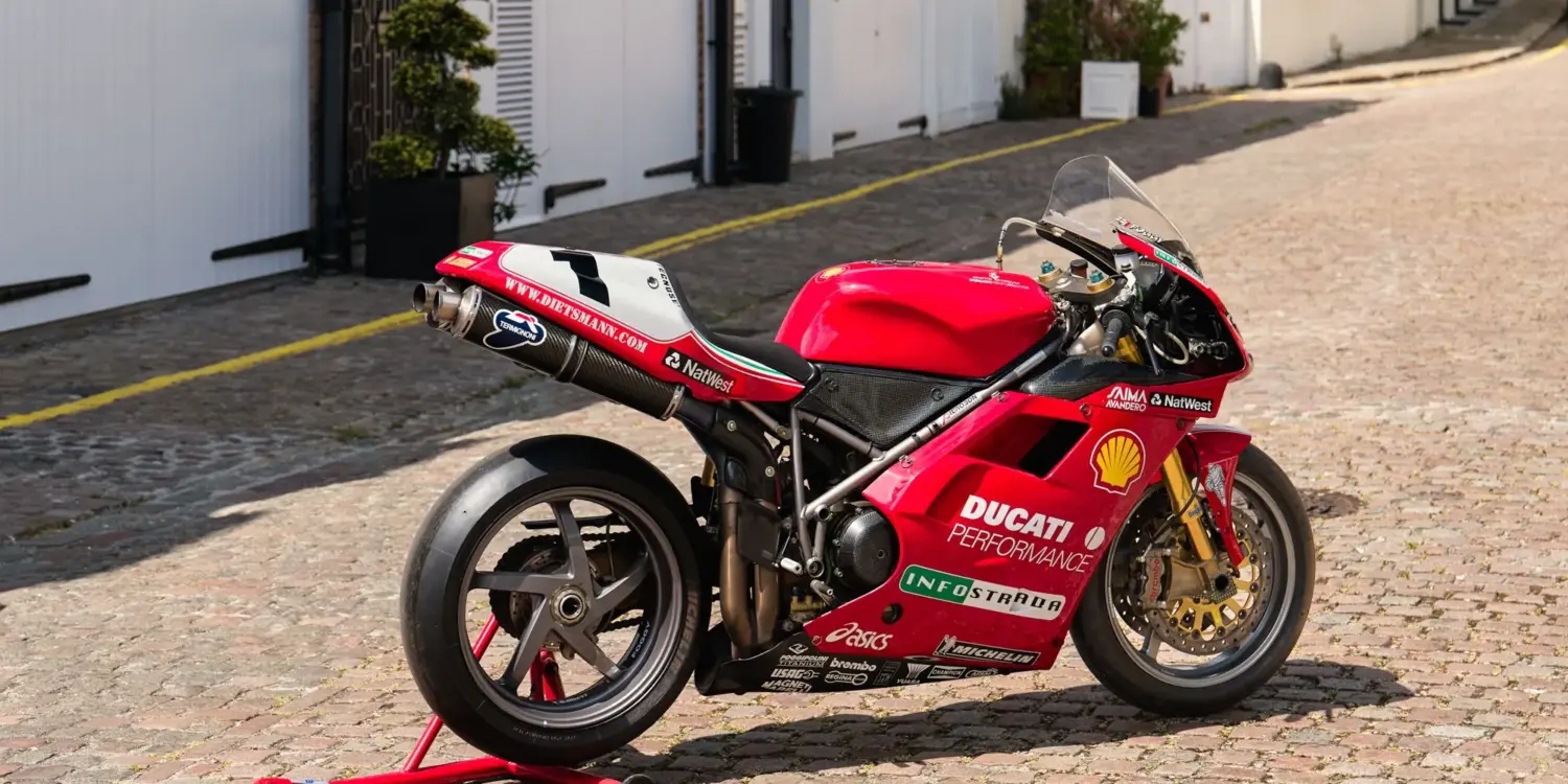 Rote Ducati 996 F99 aus 1999 mit Rennverkleidung und Sponsorenaufklebern auf einem Kopfsteinpflasterweg.