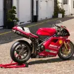 Rote Ducati 996 F99 aus 1999 mit Rennverkleidung und Sponsorenaufklebern auf einem Kopfsteinpflasterweg.