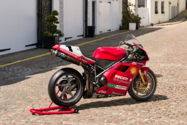 Rote Ducati 996 F99 aus 1999 mit Rennverkleidung und Sponsorenaufklebern auf einem Kopfsteinpflasterweg.