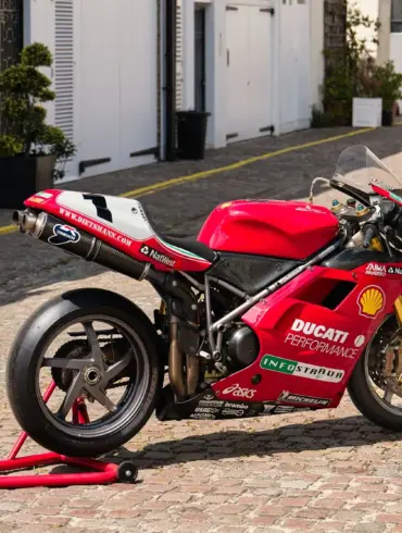 Rote Ducati 996 F99 aus 1999 mit Rennverkleidung und Sponsorenaufklebern auf einem Kopfsteinpflasterweg.