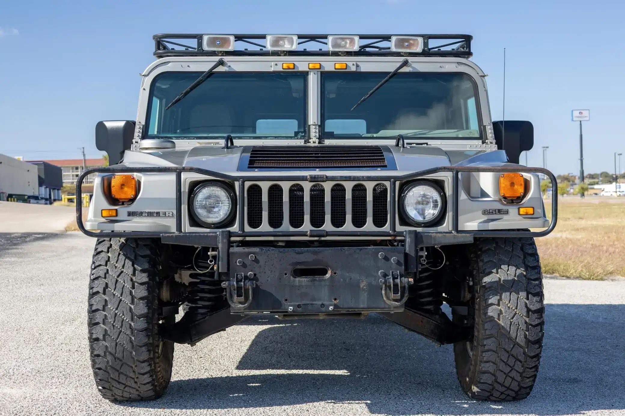 Frontansicht eines silbernen Hummer H1 Wagon aus dem Jahr 2002 auf einem Parkplatz, mit Zusatzscheinwerfern und massivem Unterbodenschutz.