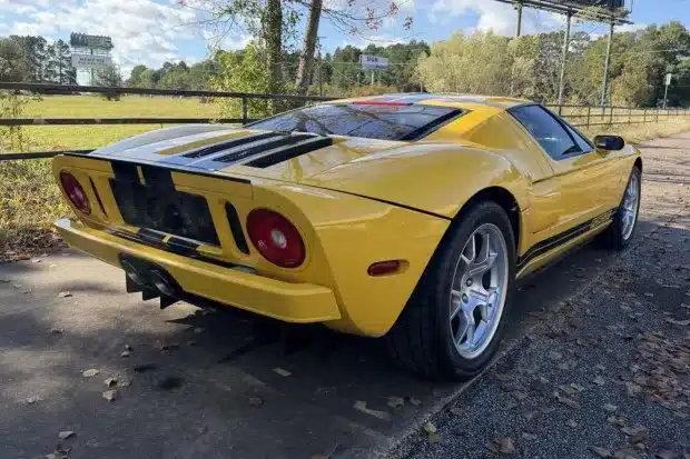 2006 Ford GT in gelber Lackierung mit schwarzen Streifen, Heckansicht auf einer Landstraße, Herbstlaub am Boden
