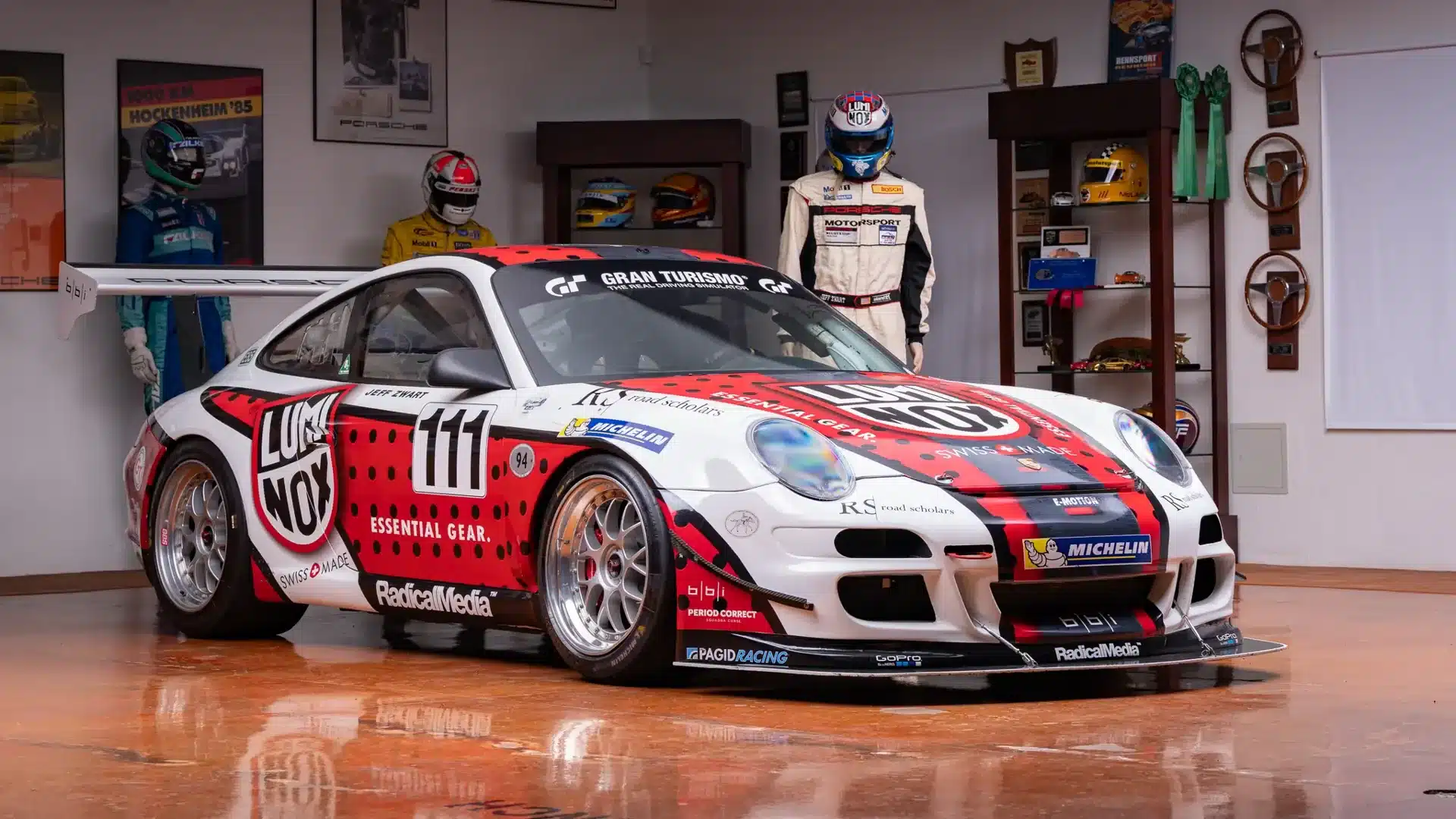 Porsche 911 GT3 Cup 2007 in Motorsportlackierung auf Hochglanzboden, umgeben von Rennanzügen, Helmen und Trophäen.