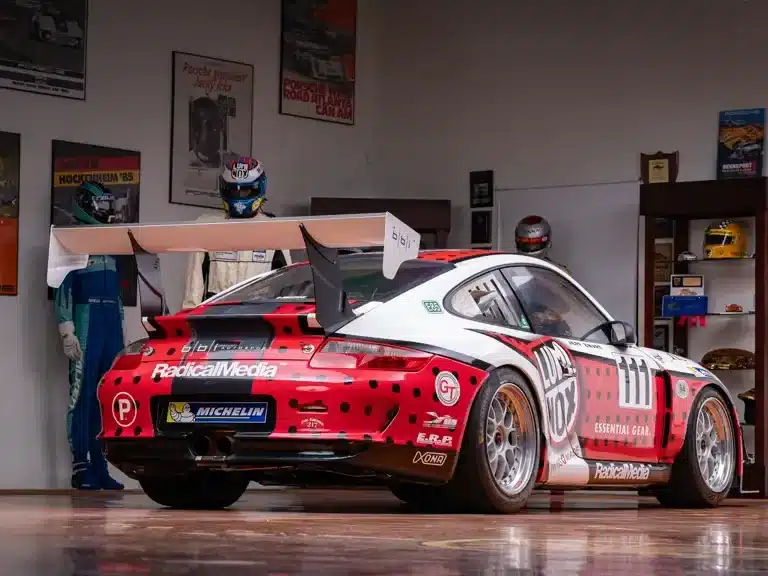 Rennwagen Porsche 911 GT3 Cup von 2007 mit auffälligem Design und großem Heckspoiler in einer Garage, umgeben von Rennfahreranzügen und Trophäen.