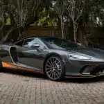 Grauer McLaren GT 2020 mit orangefarbenen Akzenten, geparkt auf einem Pflasterplatz vor Bäumen.