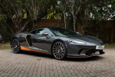 Grauer McLaren GT 2020 mit orangefarbenen Akzenten, geparkt auf einem Pflasterplatz vor Bäumen.
