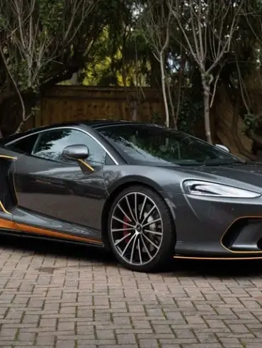 Grauer McLaren GT 2020 mit orangefarbenen Akzenten, geparkt auf einem Pflasterplatz vor Bäumen.
