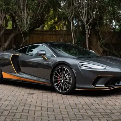 Grauer McLaren GT 2020 mit orangefarbenen Akzenten, geparkt auf einem Pflasterplatz vor Bäumen.