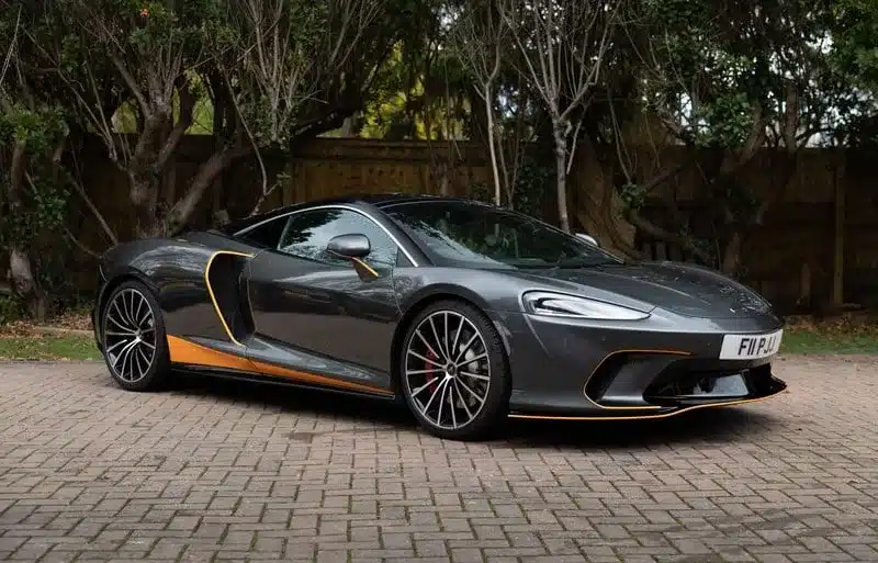 Grauer McLaren GT 2020 mit orangefarbenen Akzenten, geparkt auf einem Pflasterplatz vor Bäumen.