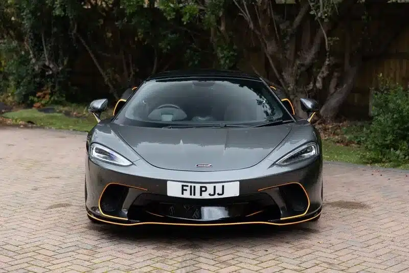 Grau lackierter McLaren GT 2020 von vorne, mit britischem Nummernschild, geparkt vor Bäumen.