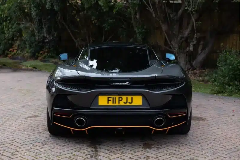 Rückansicht eines grauen McLaren GT (Baujahr 2020) mit gelbem britischem Kennzeichen, geparkt auf einer Pflasterauffahrt.