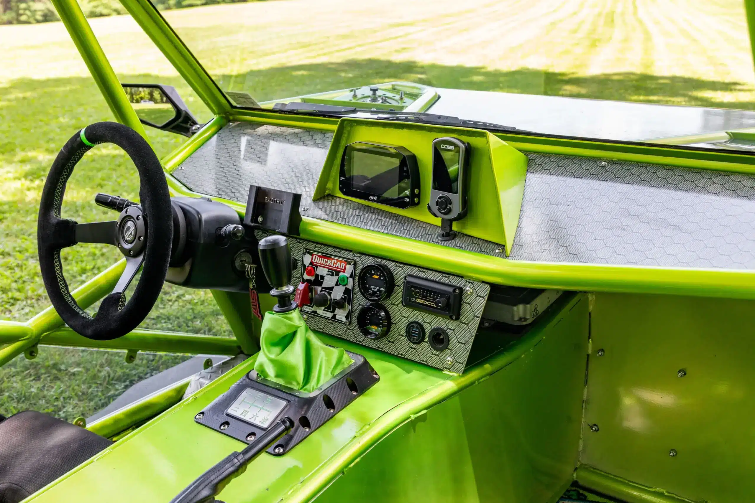 Blick auf das Cockpit eines Exotruck GR mit schwarzem NRG-Lenkrad, digitalen Anzeigen, Schaltknauf mit grüner Schaltmanschette und strukturiertem Armaturenbrett.