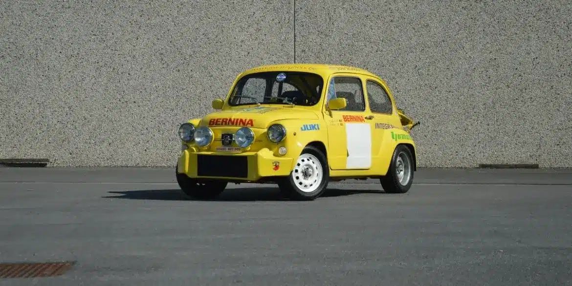 Gelber Abarth 600 1050 cm³ TC Replica mit Rennlackierung und Zusatzscheinwerfern, stehend auf Asphalt vor einer grauen Wand.