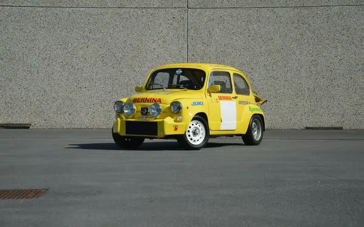 Gelber Abarth 600 1050 cm³ TC Replica mit Rennlackierung und Zusatzscheinwerfern, stehend auf Asphalt vor einer grauen Wand.