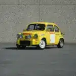 Gelber Abarth 600 1050 cm³ TC Replica mit Rennlackierung und Zusatzscheinwerfern, stehend auf Asphalt vor einer grauen Wand.