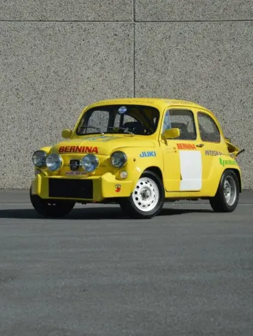 Gelber Abarth 600 1050 cm³ TC Replica mit Rennlackierung und Zusatzscheinwerfern, stehend auf Asphalt vor einer grauen Wand.