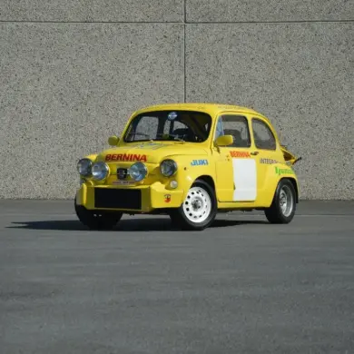 Gelber Abarth 600 1050 cm³ TC Replica mit Rennlackierung und Zusatzscheinwerfern, stehend auf Asphalt vor einer grauen Wand.