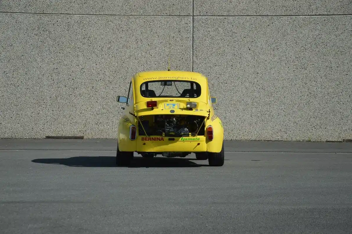 Heckansicht eines gelben Abarth 600 1050 cm³ TC Replica mit geöffnetem Motorraum auf Asphalt vor einer Wand.