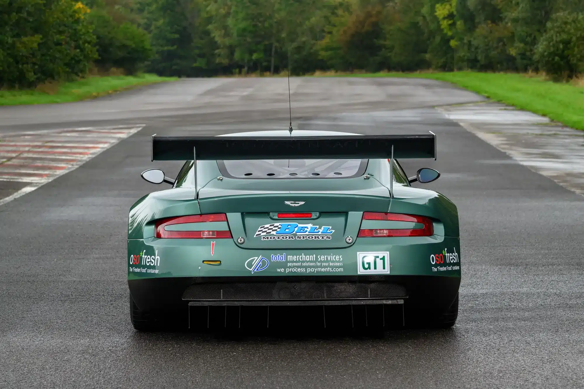 Heckansicht des grünen Aston Martin DBR9/5 GT1-Rennwagens aus dem Jahr 2006 mit großem Heckflügel und Sponsorenstickern auf einer abgesperrten Rennstrecke.