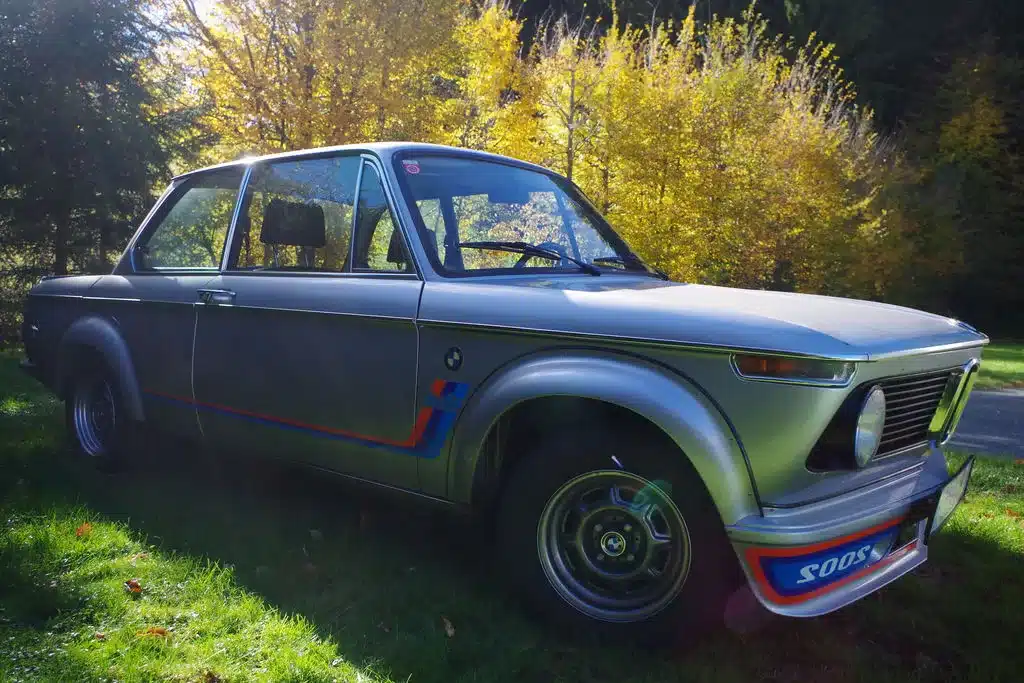 BMW 2002 Turbo von 1975 in Silber, seitlich fotografiert, im Grünen vor herbstlichen Bäumen.