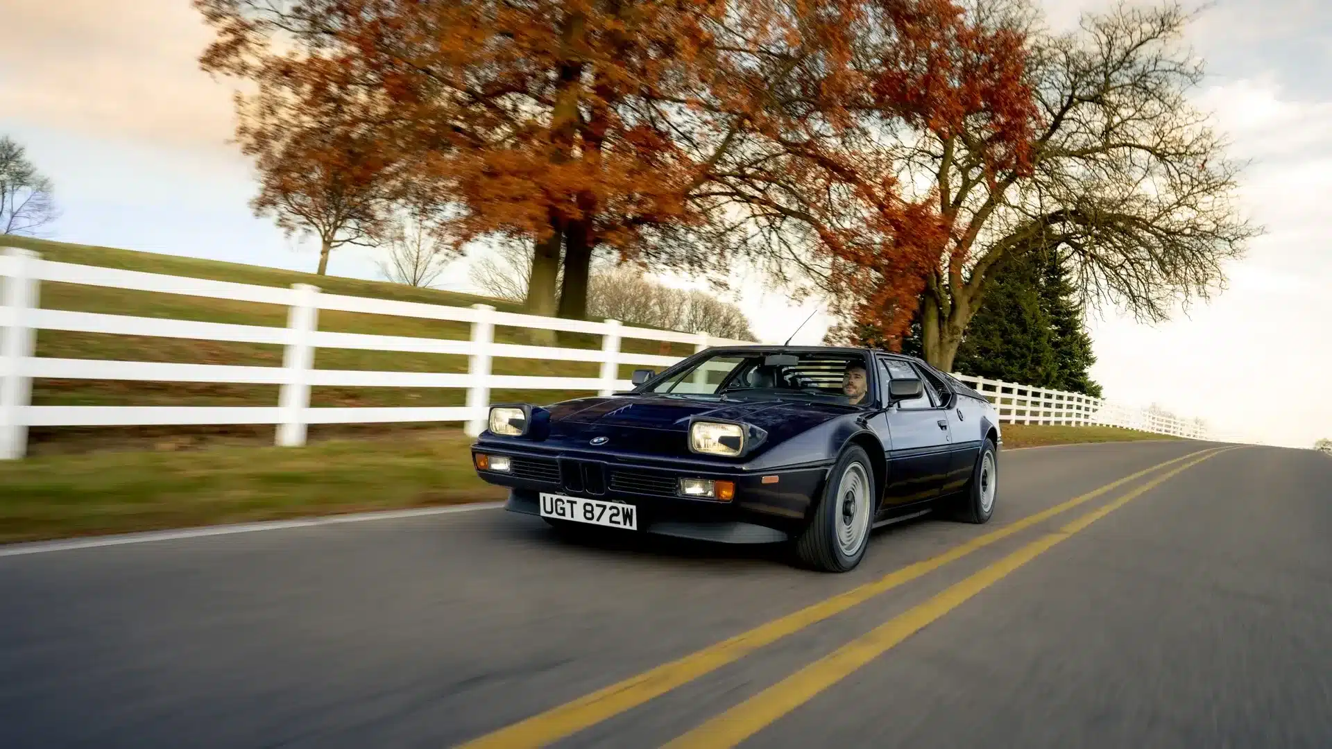 BMW M1 in dunkelblauer Lackierung fährt auf einer Landstraße mit herbstlichen Bäumen und weißem Zaun im Hintergrund.