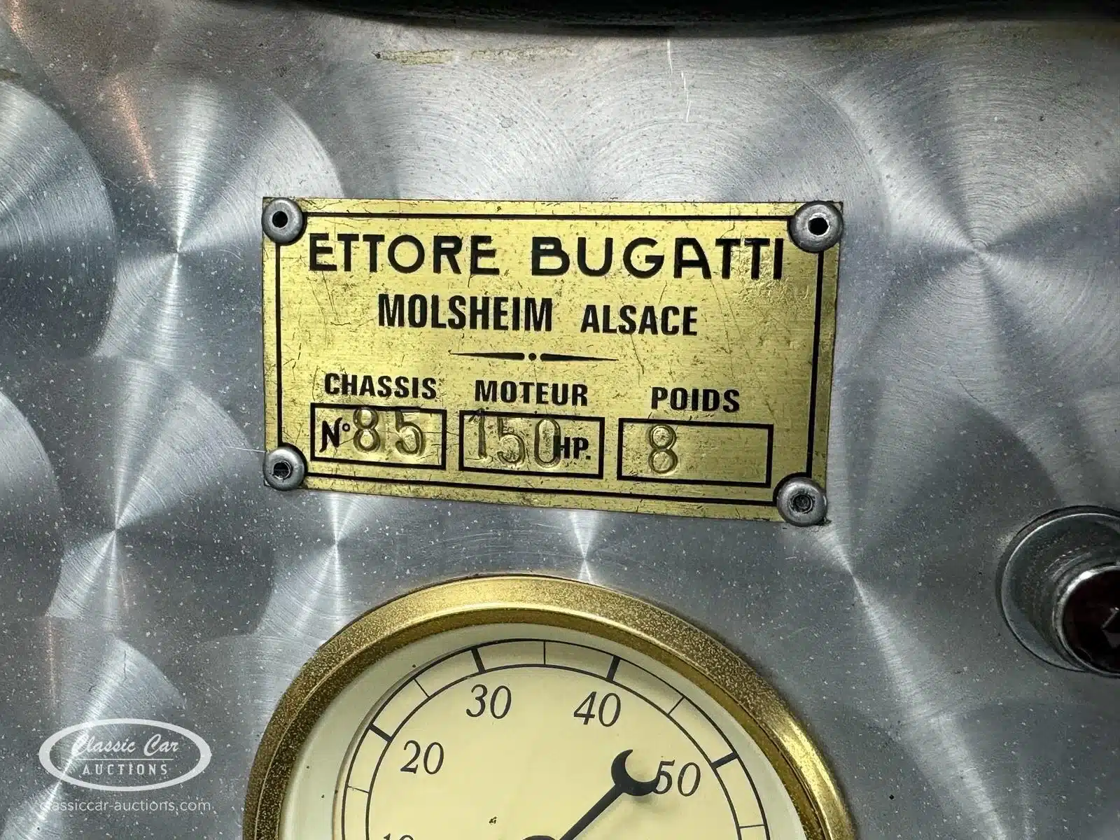 Detailansicht eines goldfarbenen Typenschilds von Ettore Bugatti mit Chassisinformationen, daneben ein klassisches Rundinstrument
