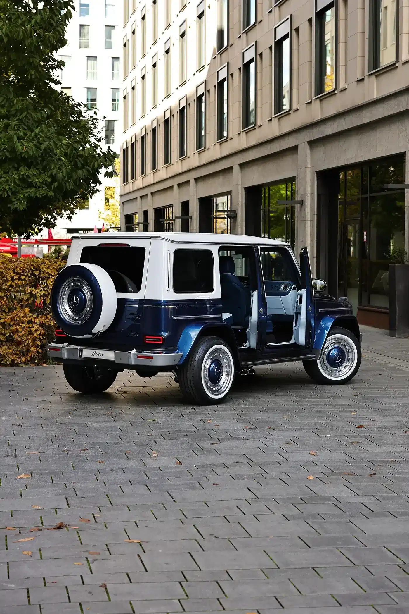 Mercedes-AMG G63 Azzurra Vintage von Carlex Design mit weiß-blauem Exterieur, individuellen Nabenkappen und geöffneter Tür in einer modernen Stadtlandschaft.