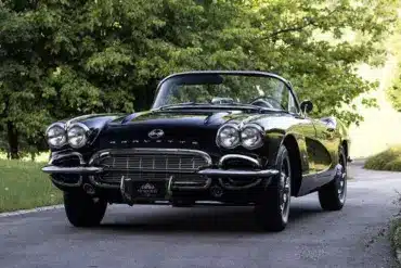 Eine schwarze Chevrolet Corvette C1 Cabriolet von 1961 steht auf einer asphaltierten Allee, umgeben von grünen Bäumen.