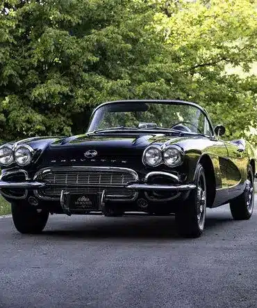 Eine schwarze Chevrolet Corvette C1 Cabriolet von 1961 steht auf einer asphaltierten Allee, umgeben von grünen Bäumen.